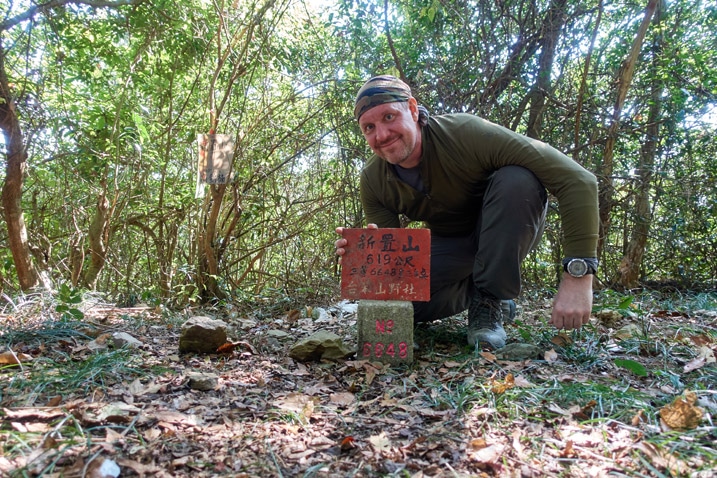 Man standing behind triangulation stone holding sign - XinZhiShan - 新置山 Peak